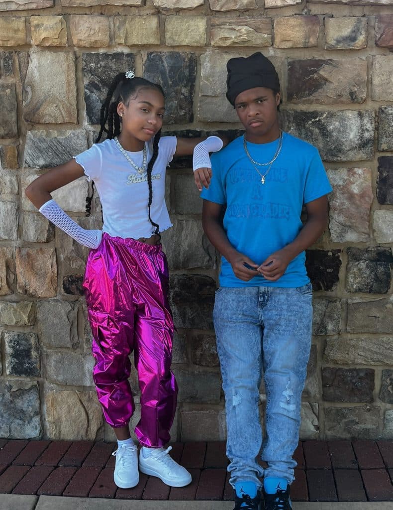 A Black female teen wearing a white top and pink pants standing with her twin brother who is wearing a blue top, jeans and dark-colored beanie.