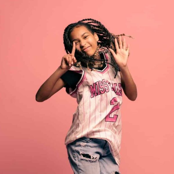 Black female teenager jumping into the air with a happy expression against a pink background.