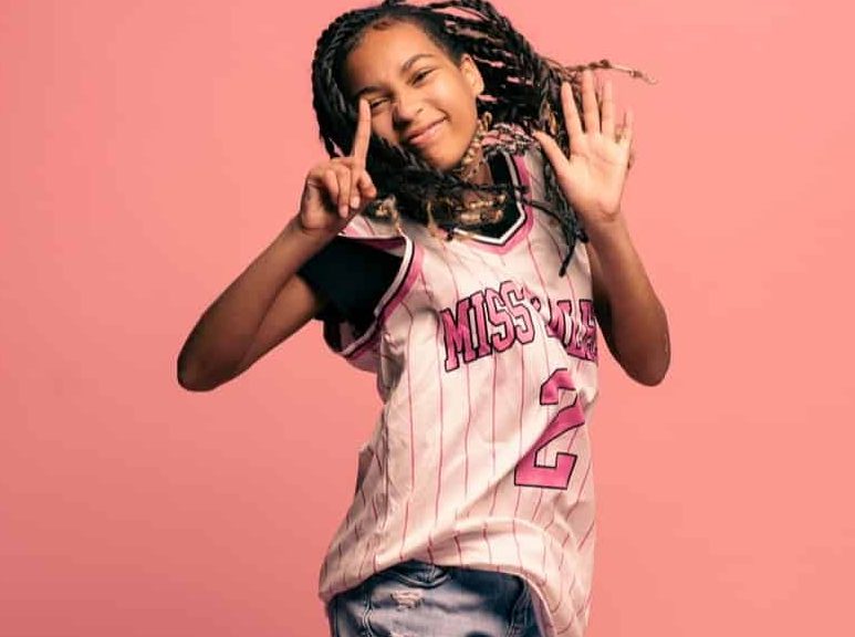 Black female teenager jumping into the air with a happy expression against a pink background.