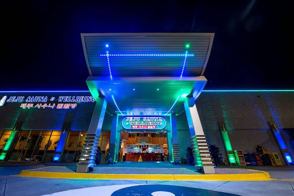 Exterior of Jeju Sauna at night, with building lit up with neon blue and green lights, set against the dark sky