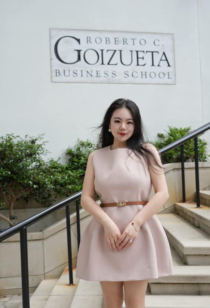 Attractive young Asian woman wearing a short, light-pink belted dress, standing on the steps outside the Goizueta Business School. Stair railings, steps and green shrubbery is behind her.