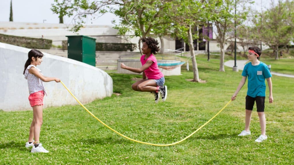 Three kids playing outside on green grass. Two are holding the ends of a long jump rope, the third is in the middle, jumping up.