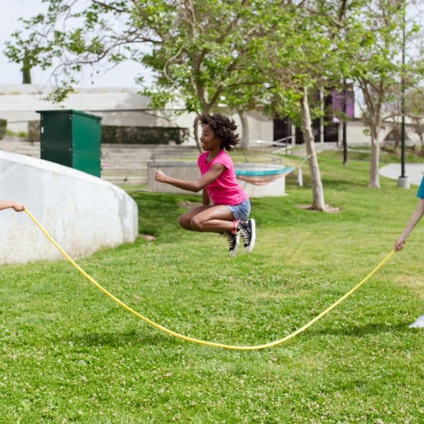 Three kids playing outside on green grass. Two are holding the ends of a long jump rope, the third is in the middle, jumping up.