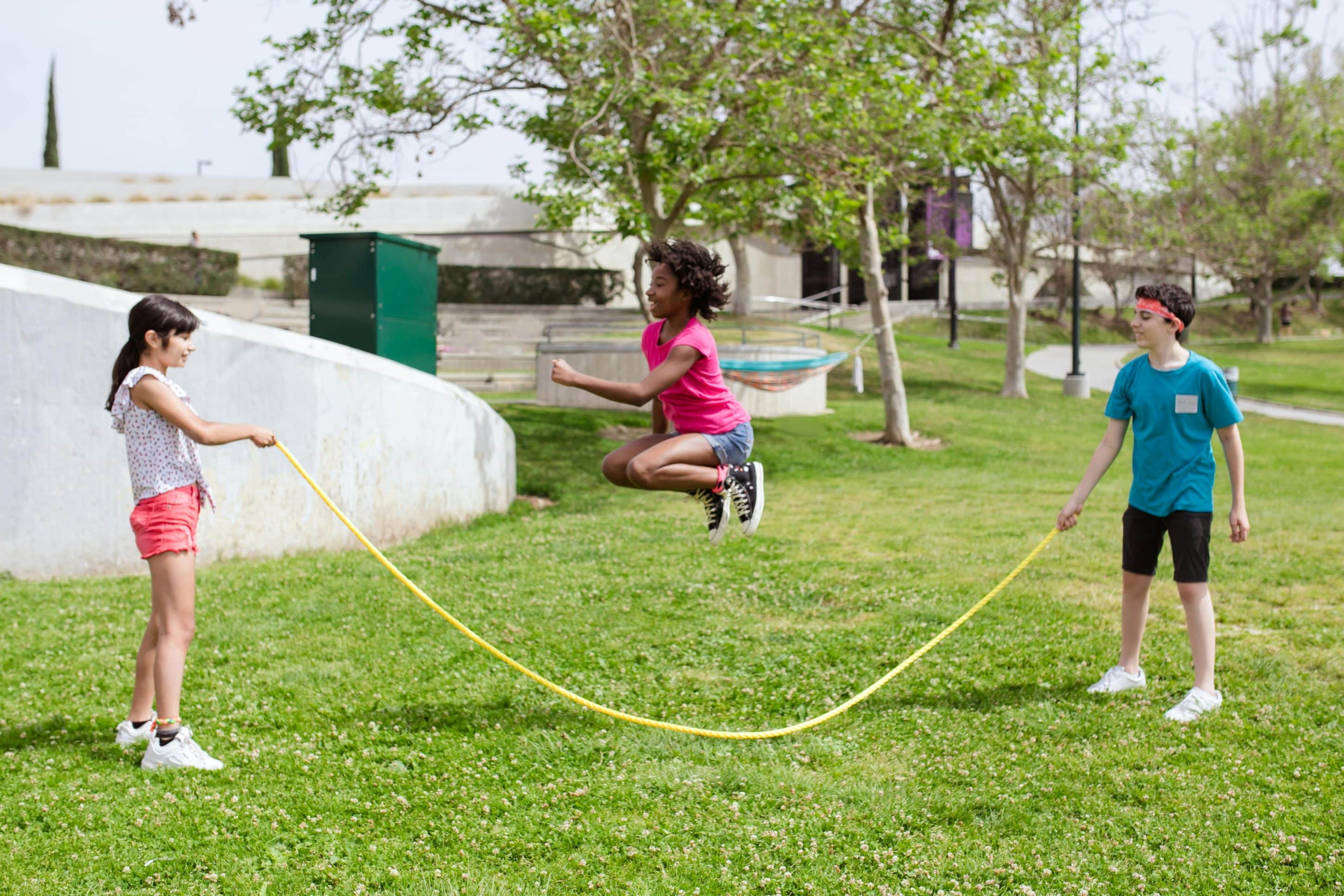 Three kids playing outside on green grass. Two are holding the ends of a long jump rope, the third is in the middle, jumping up.