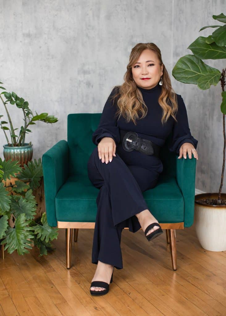 Attractive woman with long brown hair, wearing all black. She's sitting in a teal chair with thin wooden legs, with potted plants around her.