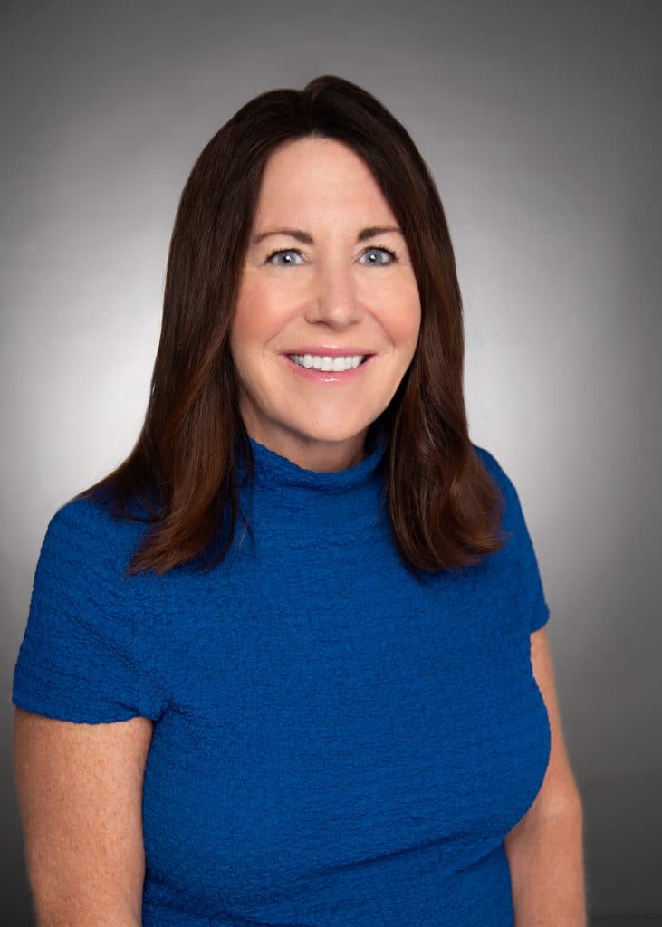 A smiling woman with shoulder-length brown hair, wearing a deep blue, short-sleeved top.