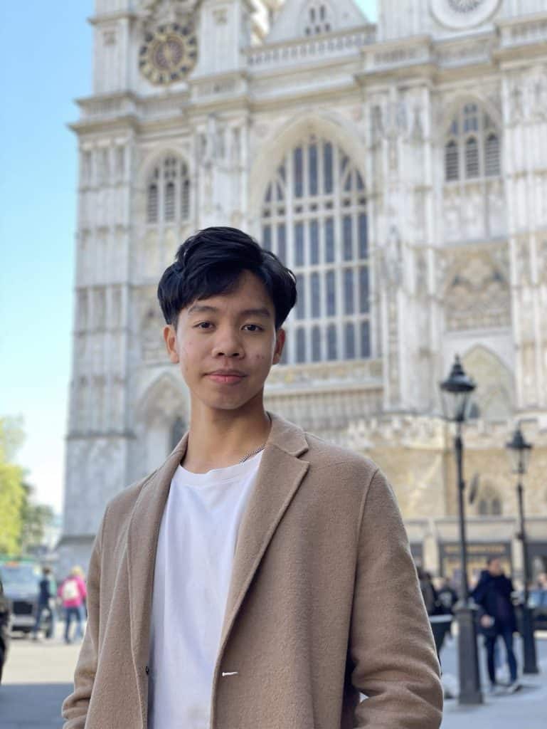 Young Asian man wearing a white tshirt and light brown jacket, standing outdoors in front of a light-stone, gothic-style building. Blue sky, green trees, light posts and people can be seen in the background