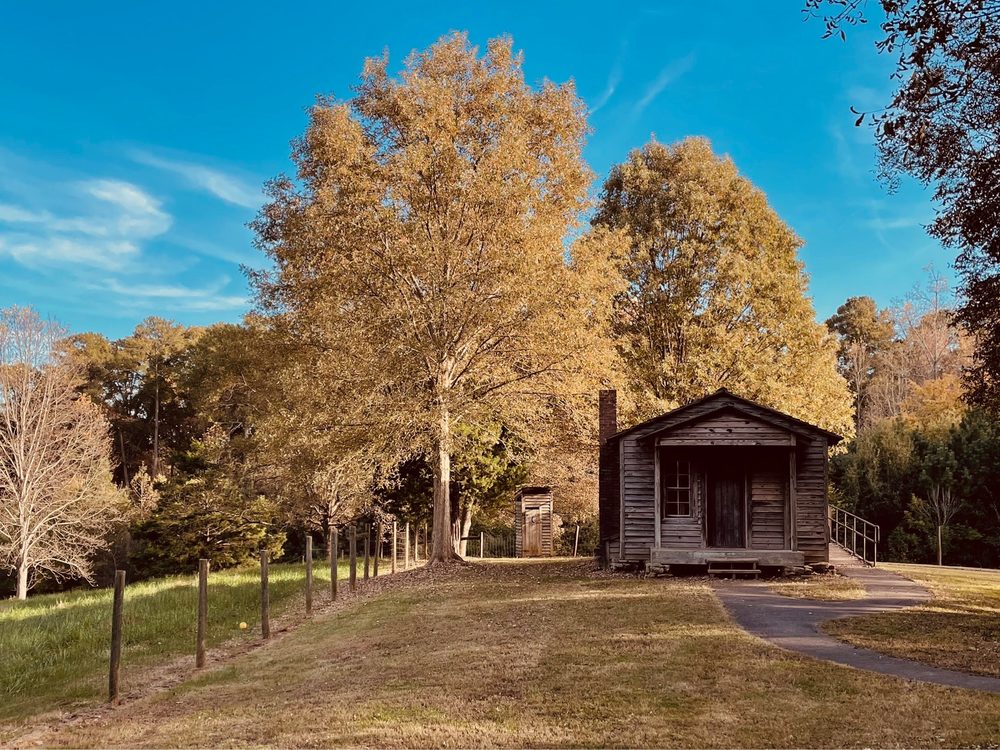 Small, historic wooden tenant home in a wooded public park. The grass is turning brown for fall and the home is surrounded by trees and line of wooden fence posts. The leaves are brown and yellow and the sky is deep blue overhead.