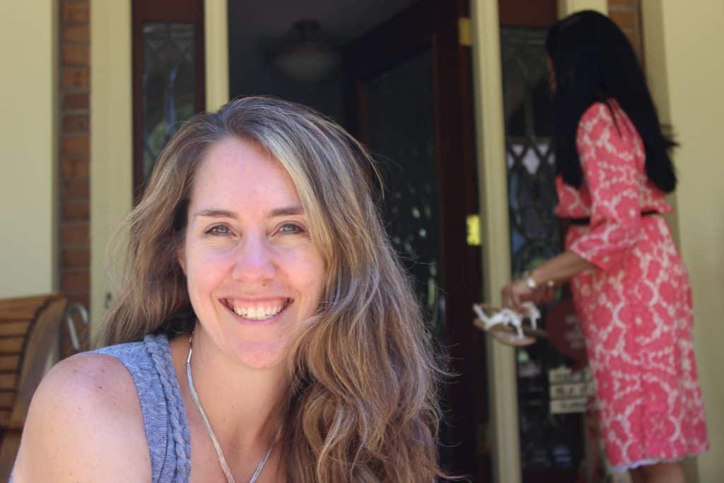 White woman with long hair, wearing a blue sleeveless top smiling at the camera. There's another woman in the background with long dark hair, wearing a red and white print dress, walking through a doorway.