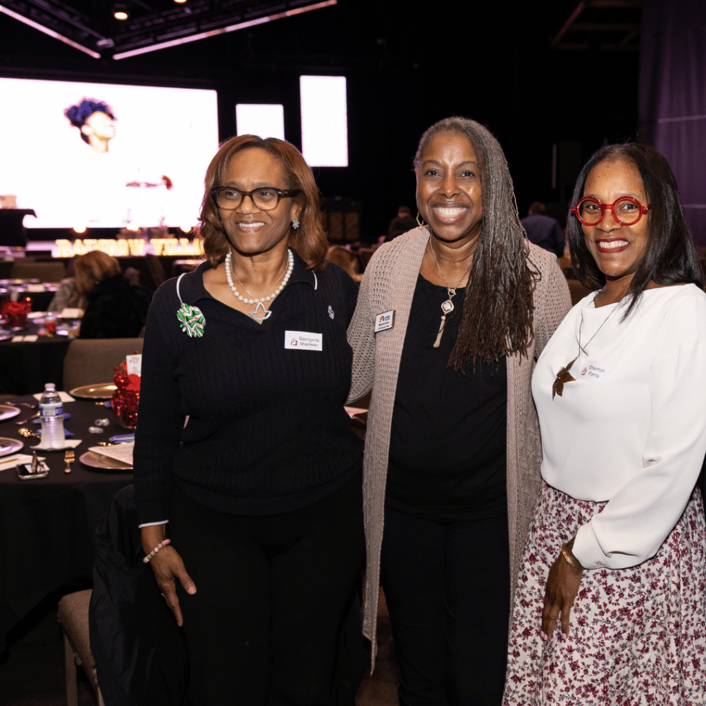 Three women, smiling and standing together for the photo, in a dimly lit conference room at a community awards event