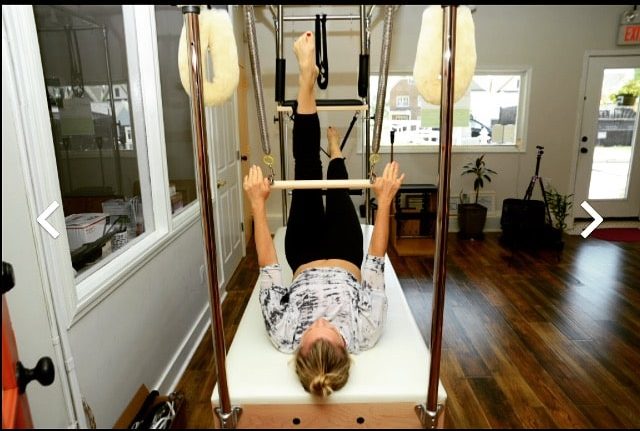 A woman with blonde hair, wearing a light colored exercise top and black leggings, lying on a Pilates machine at a small wellness center.