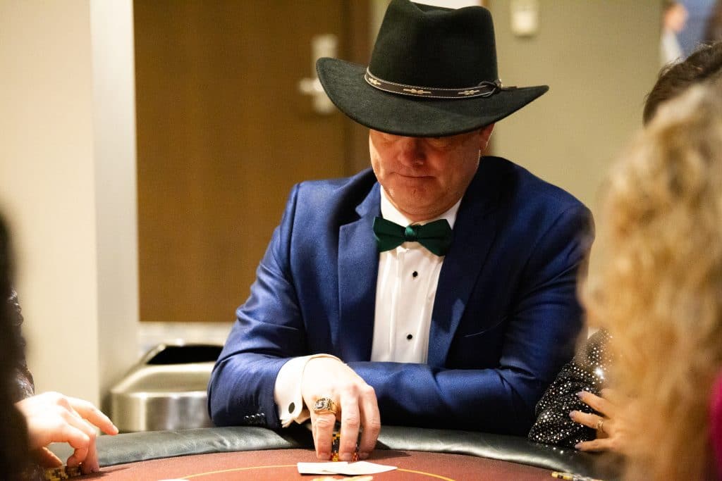 A white man in a blue suit with white shirt, wearing a wide-brimmed cowboy-style hat sitting at a card table gambling at a Las Vegas-style event.