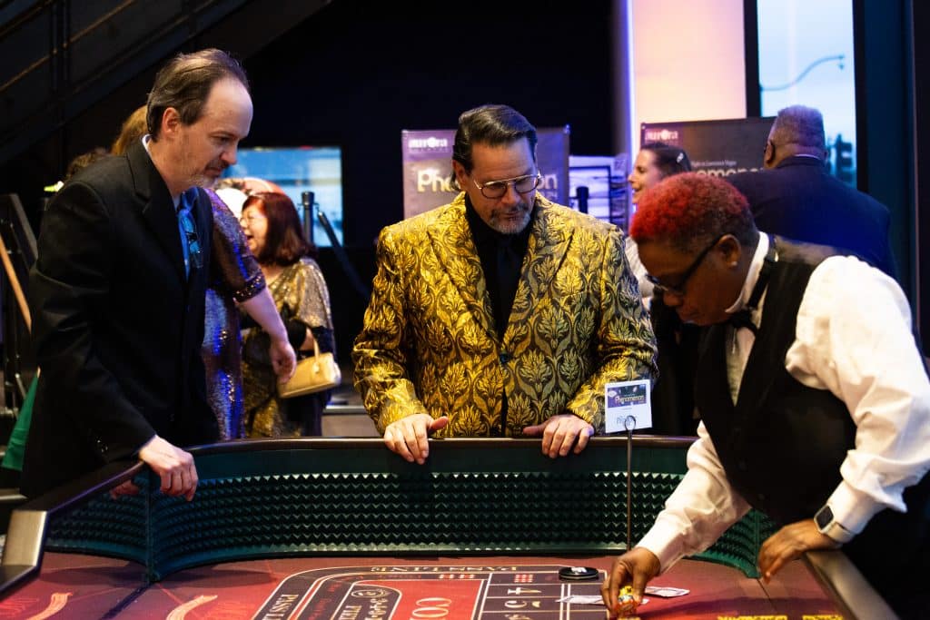 A dealer and two gamblers at a blackjack table at a Las Vegas-themed event. Other guests are in the background.