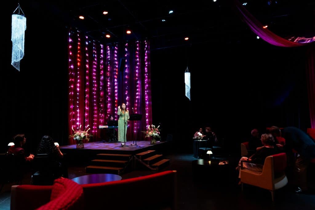 A cabaret singer onstage in a darkened room performing for the audience. People are at tables and booths watching the show. The background and lighting is a shade of deep purple.