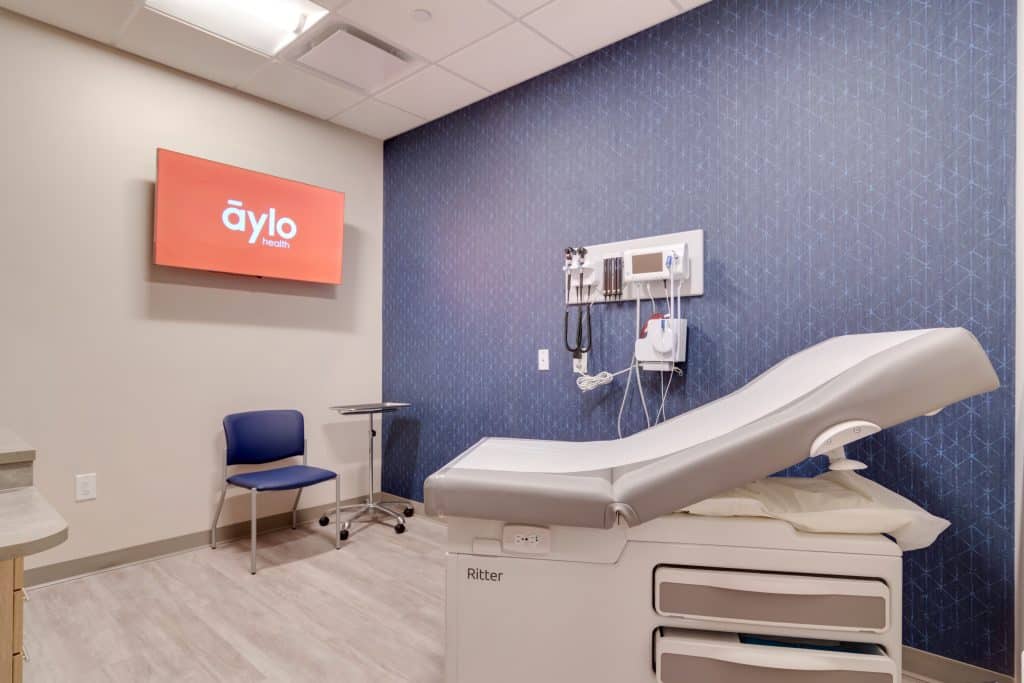 Exam room at Aylo Health in Suwanee, GA. The room is spacious with light flooring, light colored walls except for a purple accent wall, and there's an exam table, equipment and flat-screen TV/monitor.