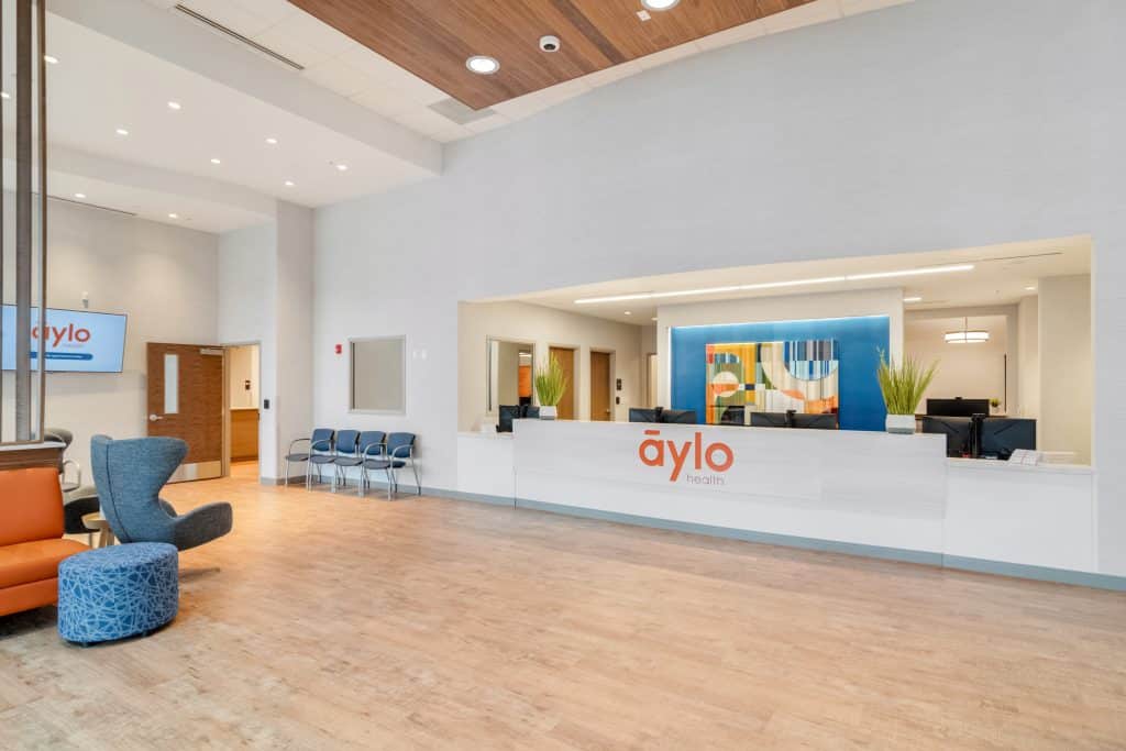 Reception and waiting room area at Aylo Health in Suwanee, GA. The space is open and modern with light wood flooring, white walls, a white built-in reception desk and different colored seats in the waiting area.