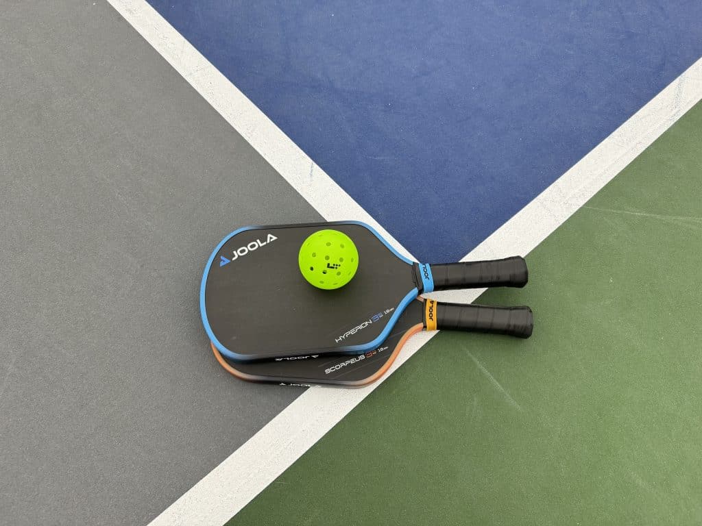 Two pickleball rackets stacked on top of each other with a green pickleball on top, resting on a pickleball court