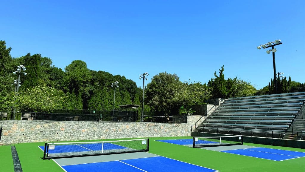 Outdoor pickleball courts with stadium seating. A row of green trees is in the background.