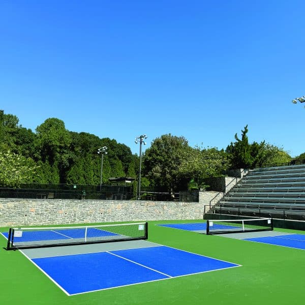 Outdoor pickleball courts with stadium seating. A row of green trees is in the background.