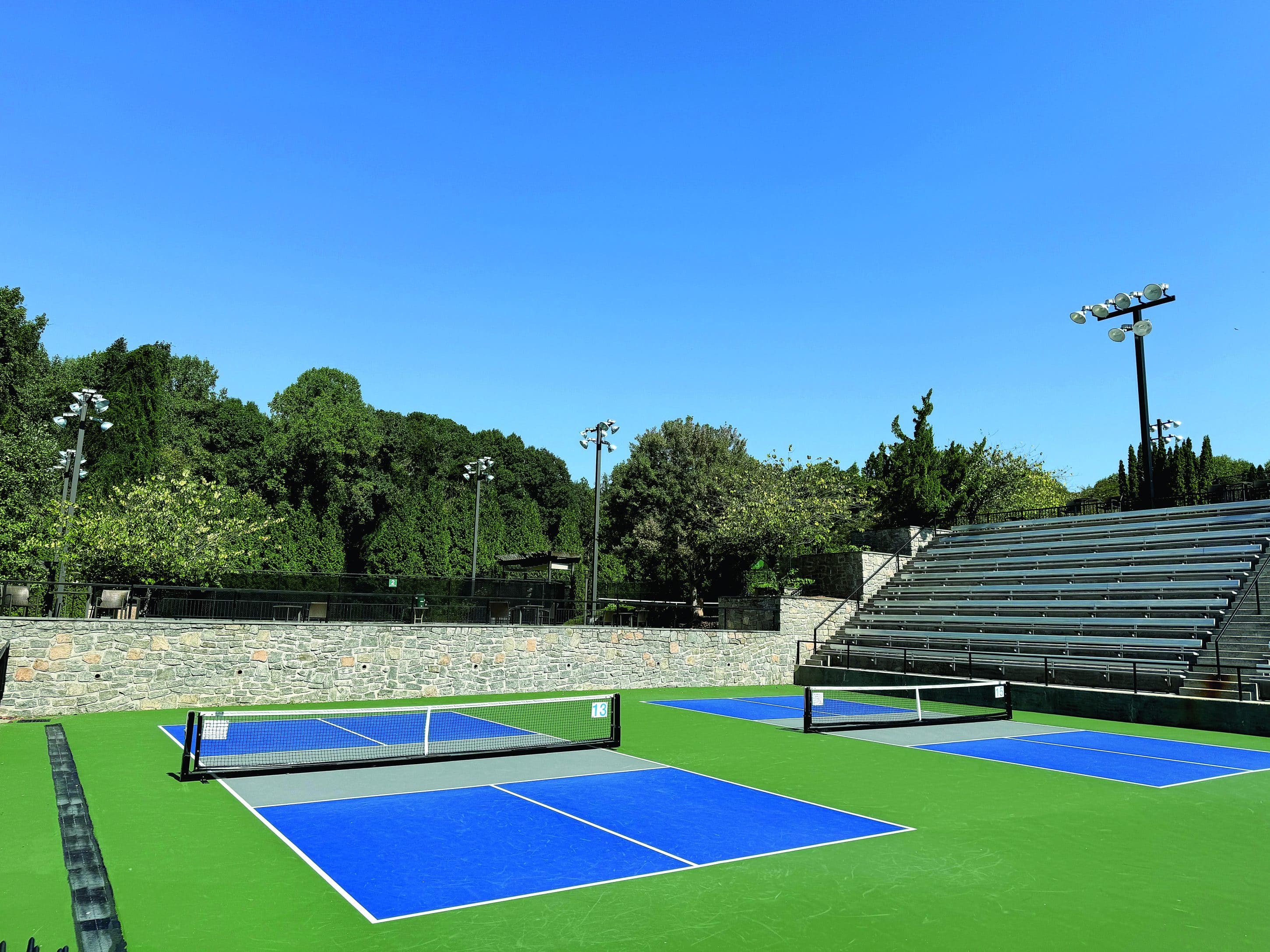 Outdoor pickleball courts with stadium seating. A row of green trees is in the background.