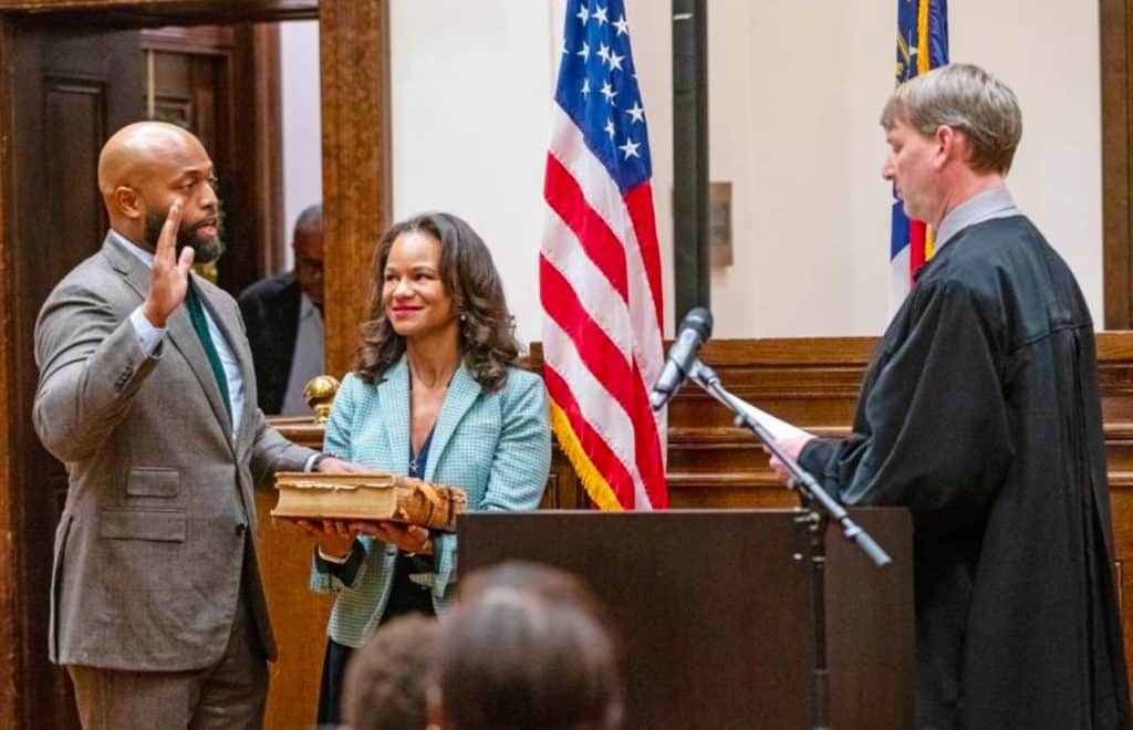 Kirkland Carden being sworn in at the Gwinnett County Commission. He's wearing a grey suit. His wife is beside him holding a Bible, a judge is in front of him and there's an American flag in the background.