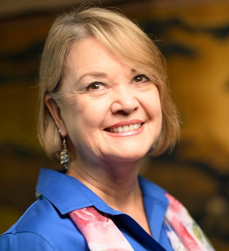 Middle-aged woman with chin-length blonde hair smiling at the camera. She's wearing a blue blouse with a collar and a pink and white flowered scarf.