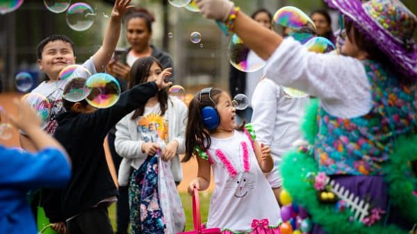 A group of families and children at an outdoor festivity. There are big bubbles being blown, everyone is smiling and laughing, and some of the kids are wearing noise-canceling headphones for sensory sensitivity.