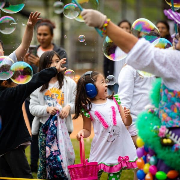 A group of families and children at an outdoor festivity. There are big bubbles being blown, everyone is smiling and laughing, and some of the kids are wearing noise-canceling headphones for sensory sensitivity.