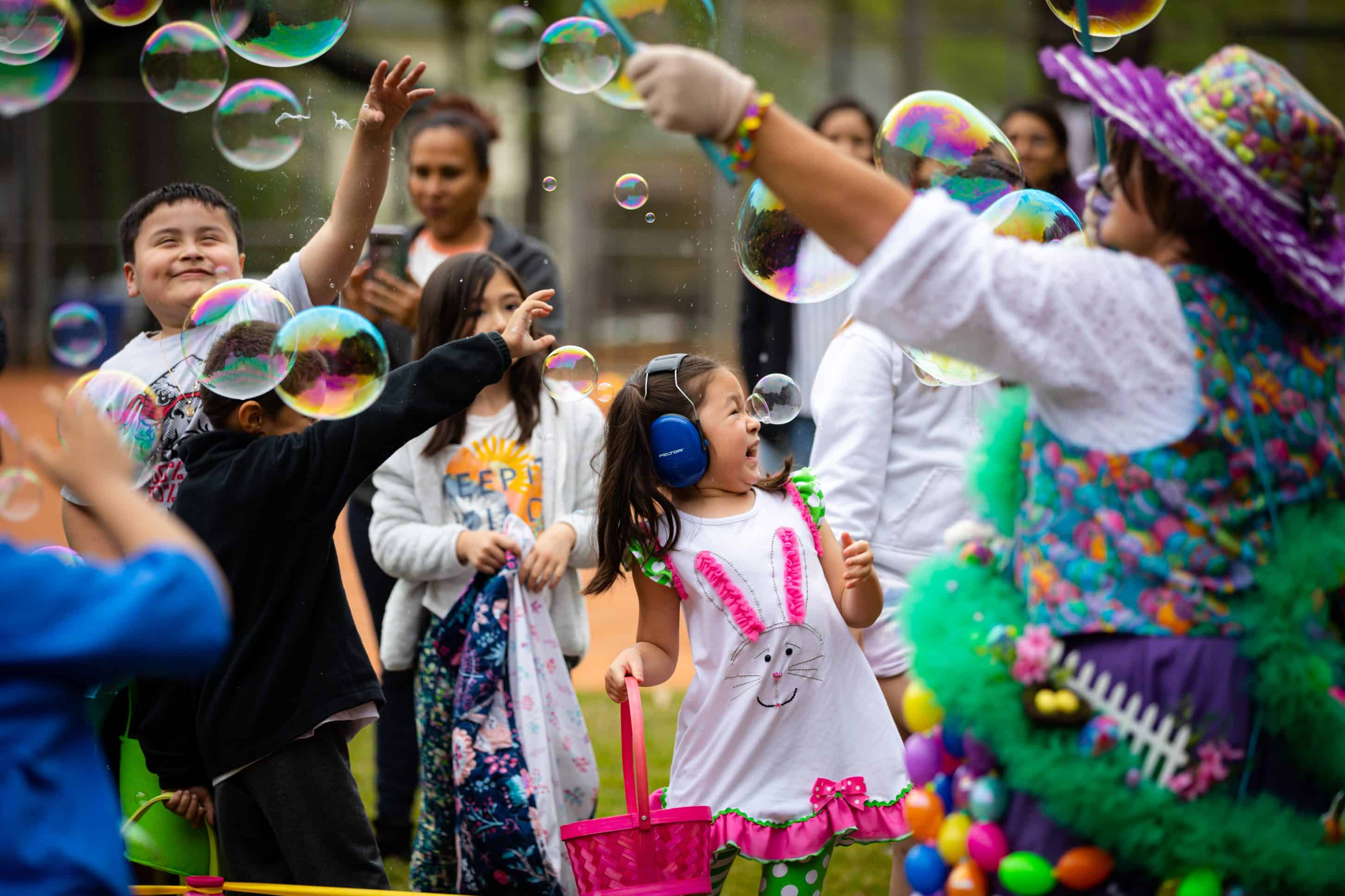 A group of families and children at an outdoor festivity. There are big bubbles being blown, everyone is smiling and laughing, and some of the kids are wearing noise-canceling headphones for sensory sensitivity.