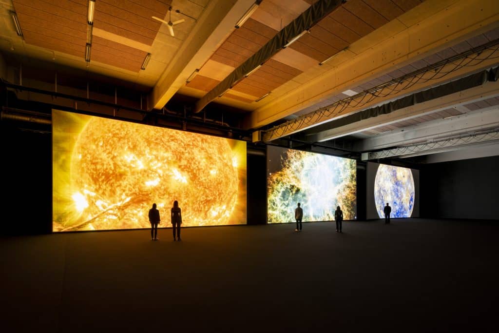 Floor-to-ceiling digital art display with three giant screens showing colorful images. a few people are silhouetted and small against the backdrop of the screens.