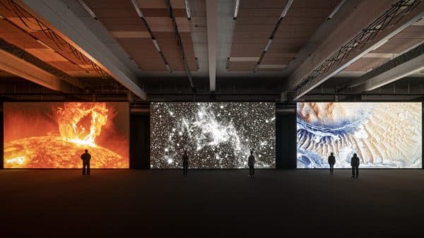 Three large, floor-to-ceiling screens showing a projected digital art exhibit from Ryoji Ikeda. A few viewers are small and silhouetted agains the screens.