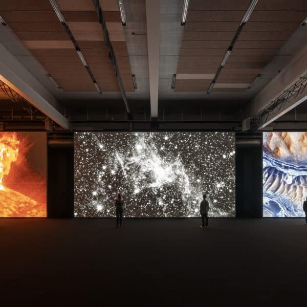 Three large, floor-to-ceiling screens showing a projected digital art exhibit from Ryoji Ikeda. A few viewers are small and silhouetted agains the screens.