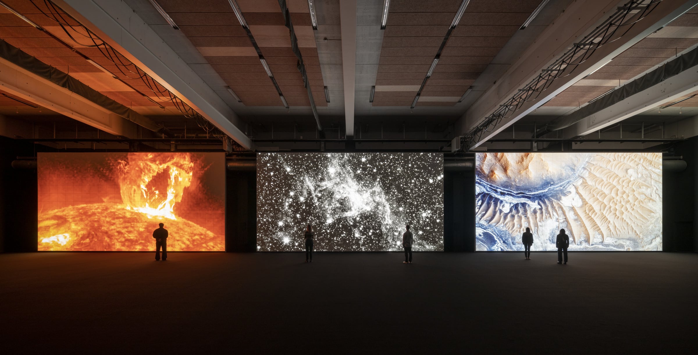 Three large, floor-to-ceiling screens showing a projected digital art exhibit from Ryoji Ikeda. A few viewers are small and silhouetted agains the screens.