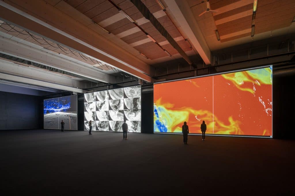 Three large, floor-to-ceiling screens showing a projected digital art exhibit from Ryoji Ikeda. A few viewers are small and silhouetted agains the screens.