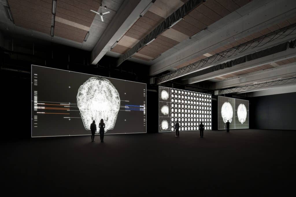 Three large, floor-to-ceiling screens showing a projected digital art exhibit from Ryoji Ikeda. A few viewers are small and silhouetted agains the screens.