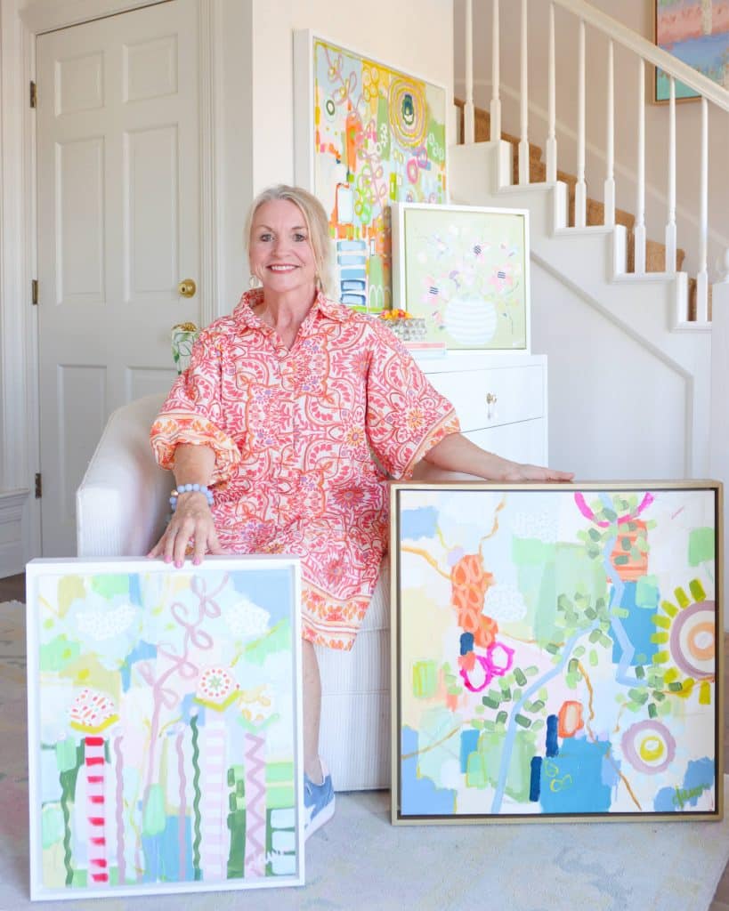 A blonde-haired woman wearing a flowing pink and white dress, standing in a light and airy room with two of her pastel abstract paintings.