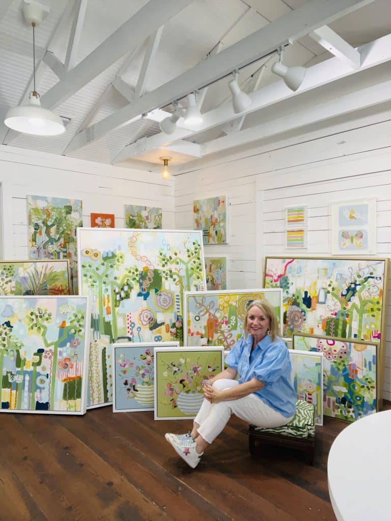 A woman with blonde hair, wearing a blue top and white pants sits on a wood floor surrounded by a dozen or so pastel-colored paintings.