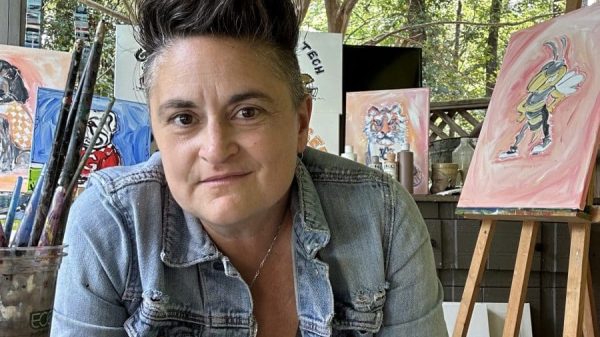 A woman with short dark hair wearing a black shirt and blue jean jacket. She's sitting on a porch with paintings behind her.