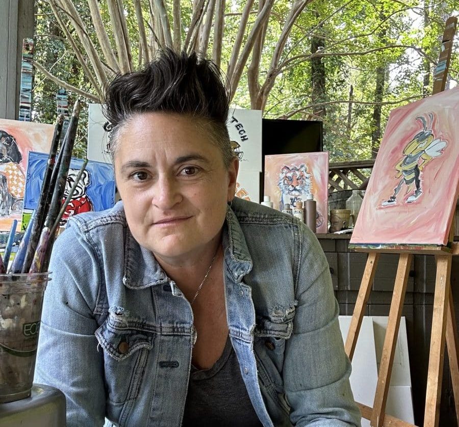 A woman with short dark hair wearing a black shirt and blue jean jacket. She's sitting on a porch with paintings behind her.