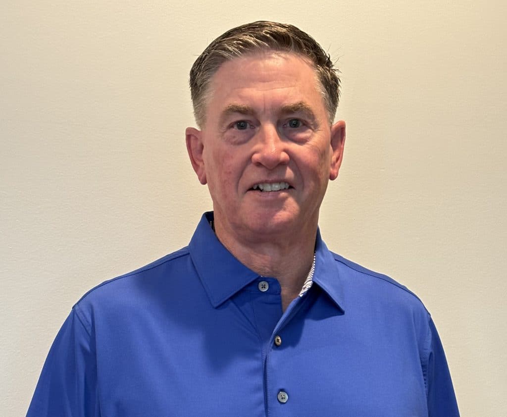 An older, middle-aged white man with short brown and grey hair. He's wearing a blue, button-up collar shirt and standing in front of a beige-colored wall.