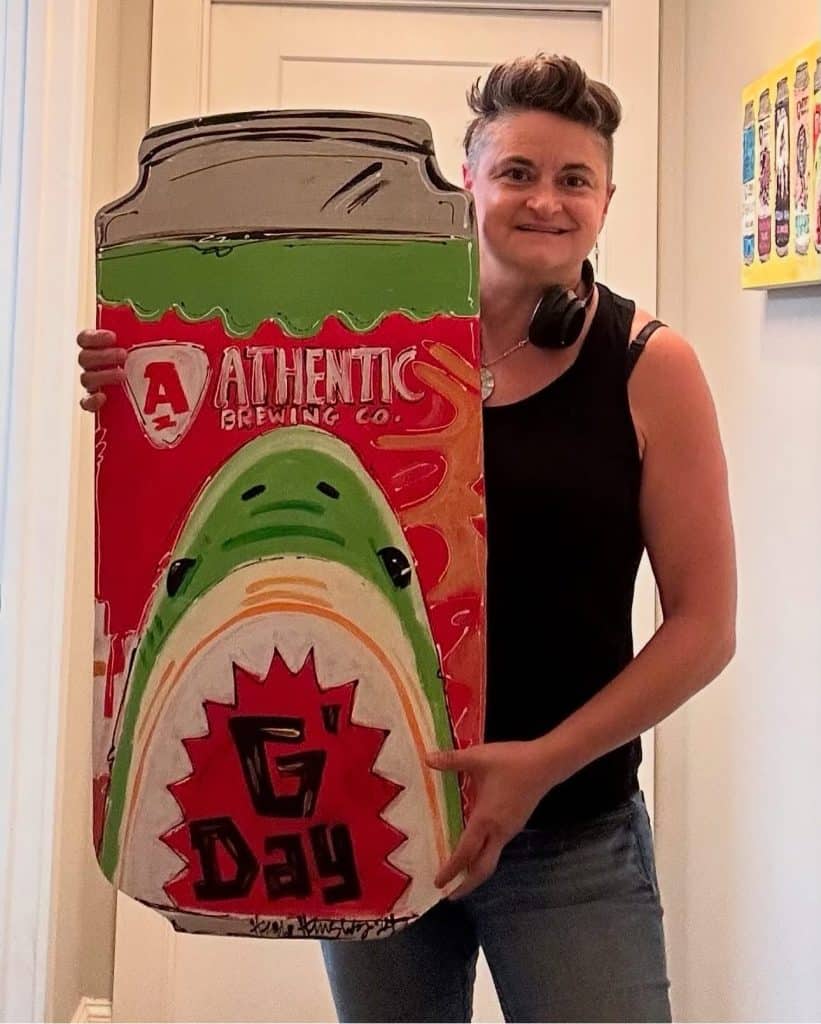 A woman with short dark hair, wearing black and holding a colorful, painted cut-out of a canned beverage