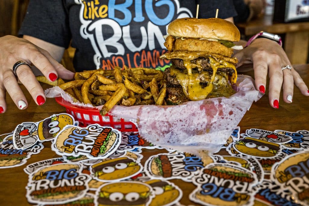 Close up of a woman's hands with red and white painted nails next to a basket with a large burger and fries. There are stickers for Gwinnett Burger Week on the table in front of her.