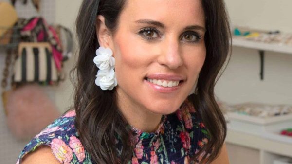 Woman with long, dark hair parted on the side. She's wearing a floral patterned sleeveless dress and large, light-colored earings.