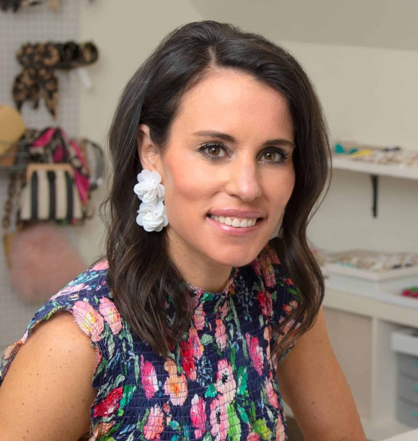 Woman with long, dark hair parted on the side. She's wearing a floral patterned sleeveless dress and large, light-colored earings.