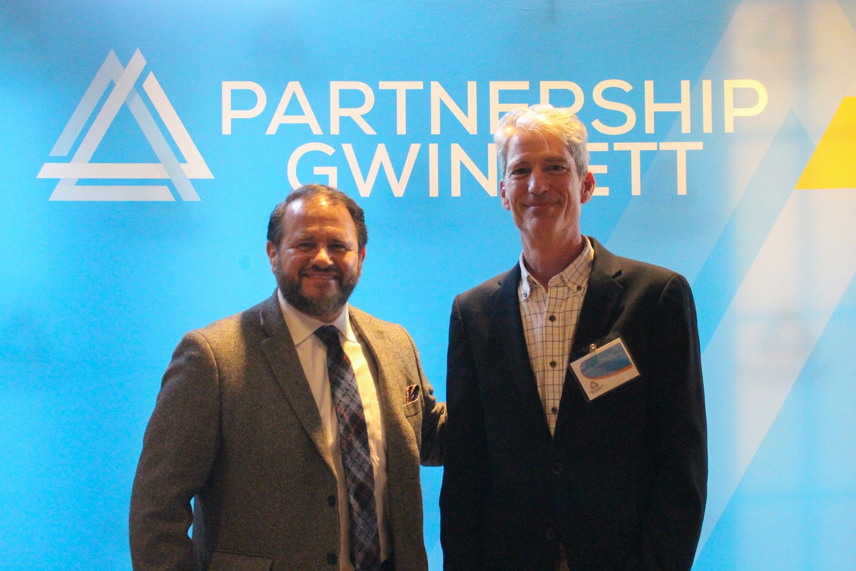 Two men in suits smiling and standing next to each other in front of Partnership Gwinnett backdrop with white lettering on blue background.