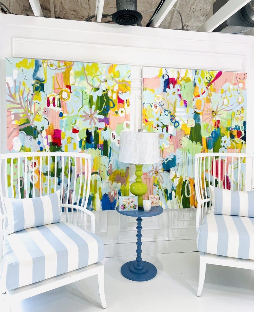 Two colorful, mixed-media abstract paintings on a wall behind two white and blue striped chairs and aw blue floorlamp with white shade.