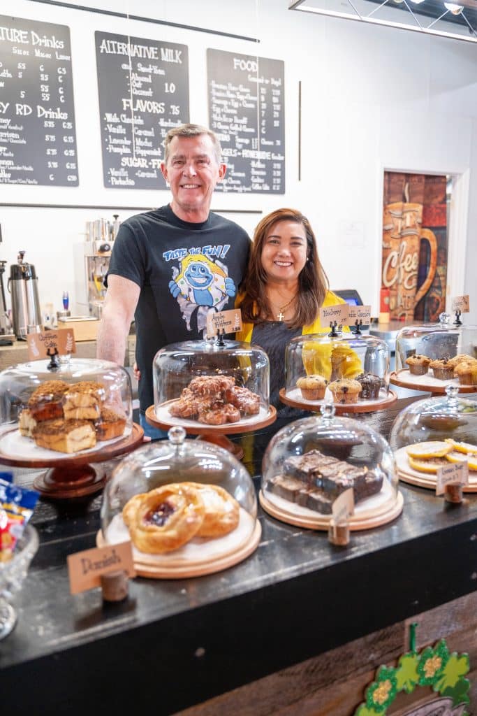 A man and woman standing together behind a counter. In front of them are displays of homemade pastries and baked goods.