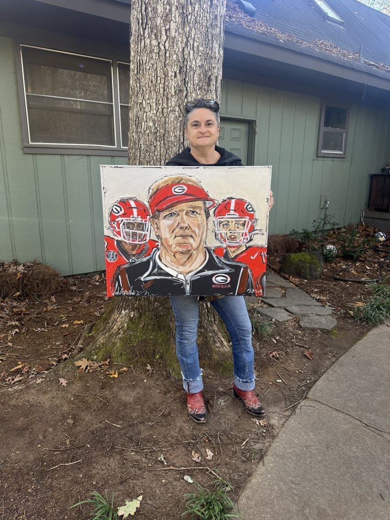 A woman with short dark hair standing outside next to a tree holding a UGA painting