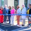 A group of people standing in front of a health clinic holding a long blue ribbon with Good Samaritan Health Center written on it. They are preparing to cut the ribbon to open the new center.