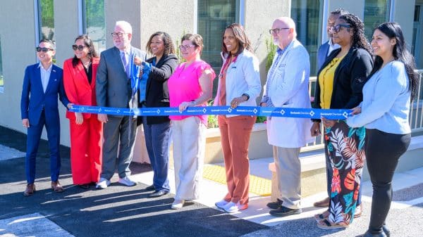 A group of people standing in front of a health clinic holding a long blue ribbon with Good Samaritan Health Center written on it. They are preparing to cut the ribbon to open the new center.
